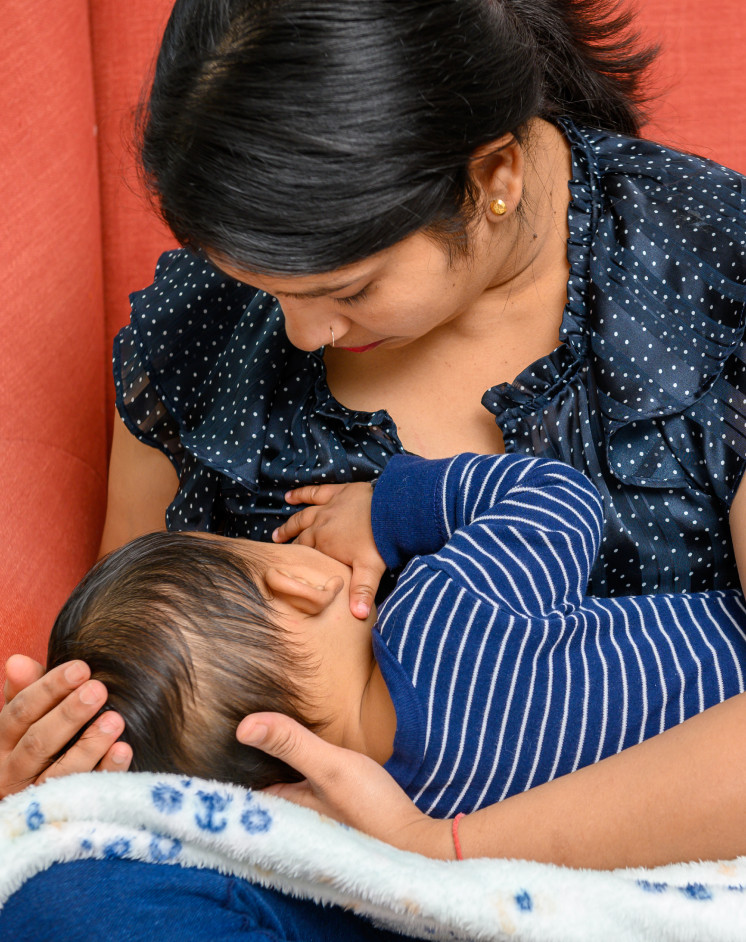 Một người phụ nữ mặc áo sơ mi chấm bi màu xanh hải quân đang cho một em bé mặc bộ đồ sọc xanh bú sữa mẹ, trong khi ngồi trên một chiếc ghế màu cam.