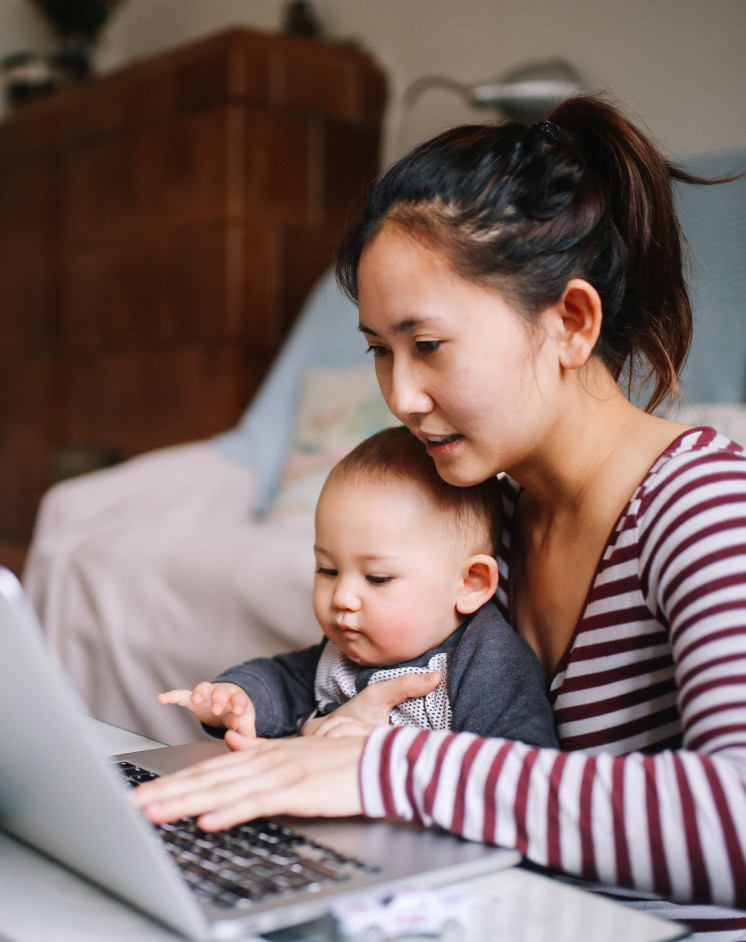 Một người phụ nữ ngồi với một em bé trên đùi, cả hai cùng nhìn vào màn hình máy tính xách tay và gõ bàn phím.
